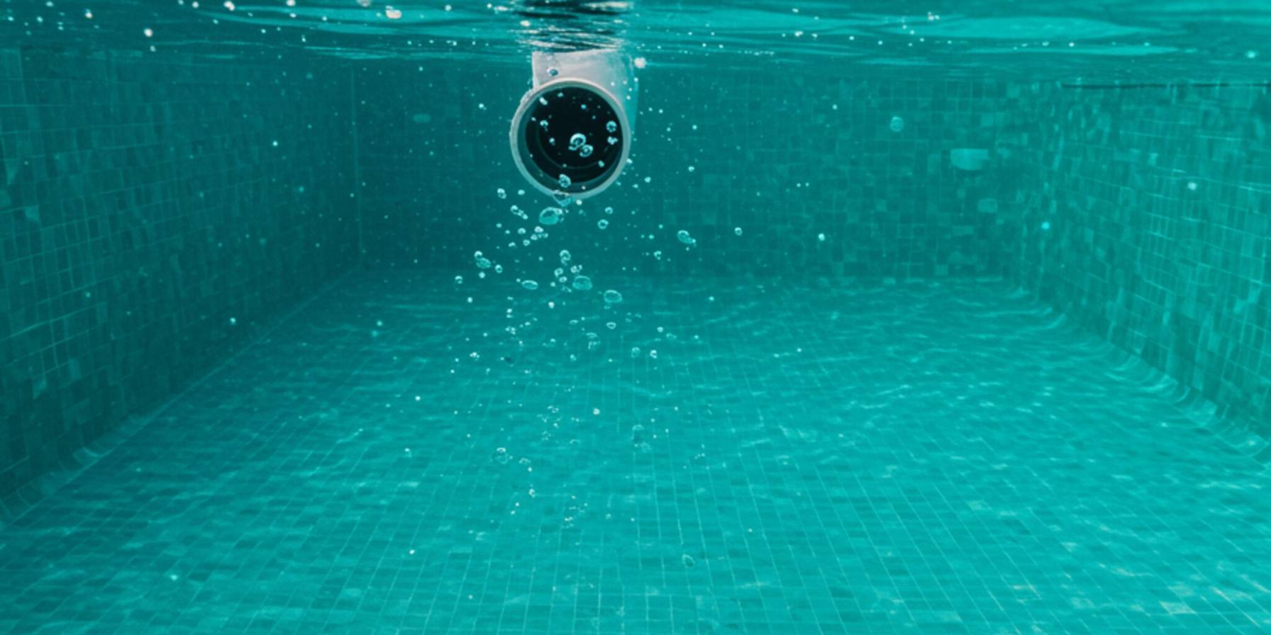 "Perspectiva submarina de una piscina de lujo en una villa andaluza, mostrando burbujas de aire de una fuga en una unión de tubería, mientras un técnico utiliza una cámara digital para detectar fugas."