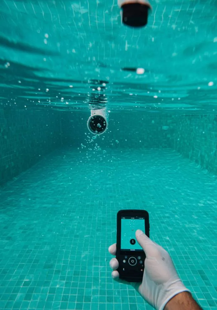 "Perspectiva submarina de una piscina de lujo en una villa andaluza, mostrando burbujas de aire de una fuga en una unión de tubería, mientras un técnico utiliza una cámara digital para detectar fugas."
